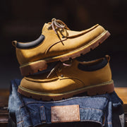 A pair of tan suede lace-up shoes stacked on top of folded blue jeans against a dark blurred background.