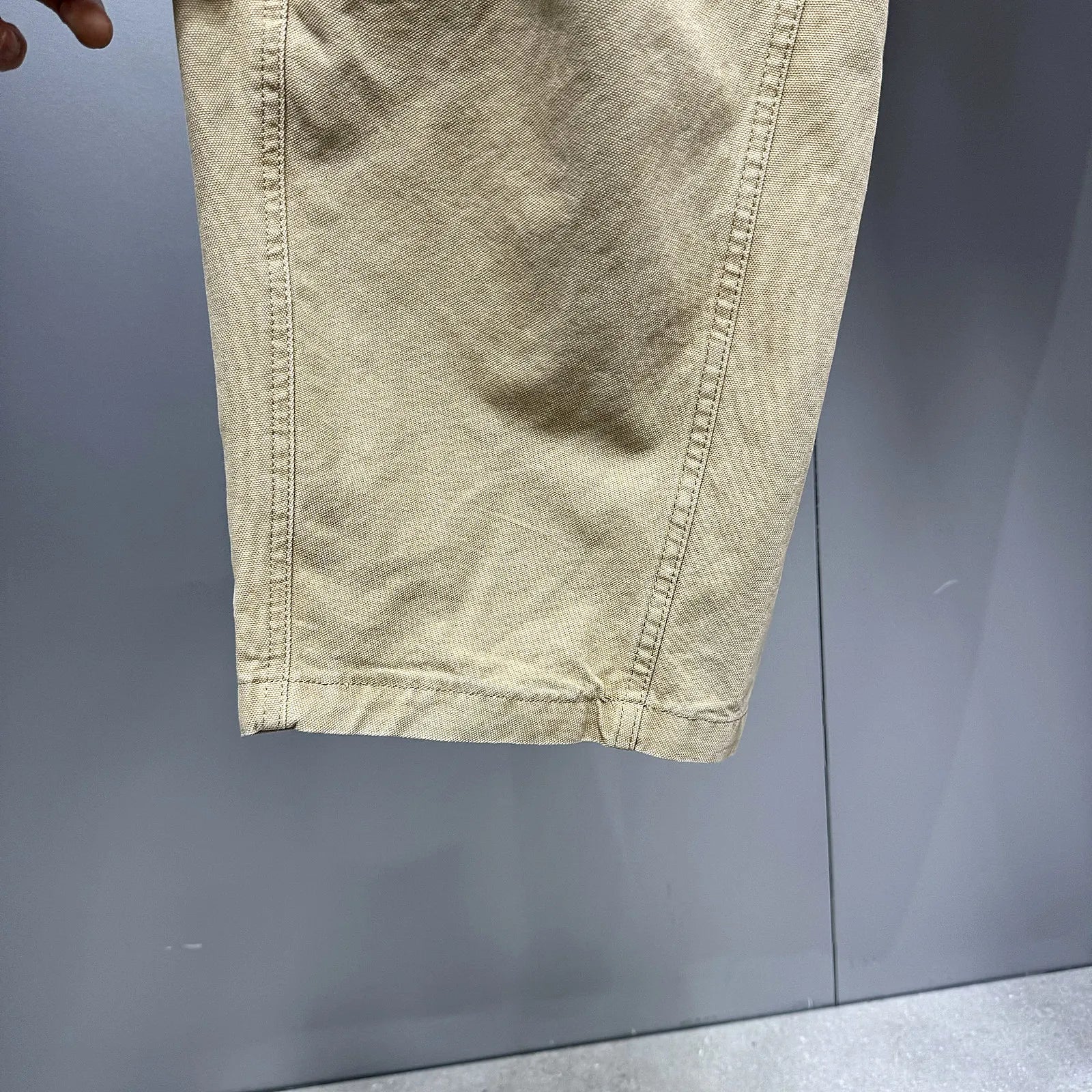 A close-up of a light beige or khaki fabric garment, showing the texture and hem stitching, held against a gray background.