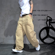 A person wearing loose beige cargo-style pants, a white sleeveless top, and white sneakers stands in a studio setting with a gray textured wall and a black bicycle prop in the background.