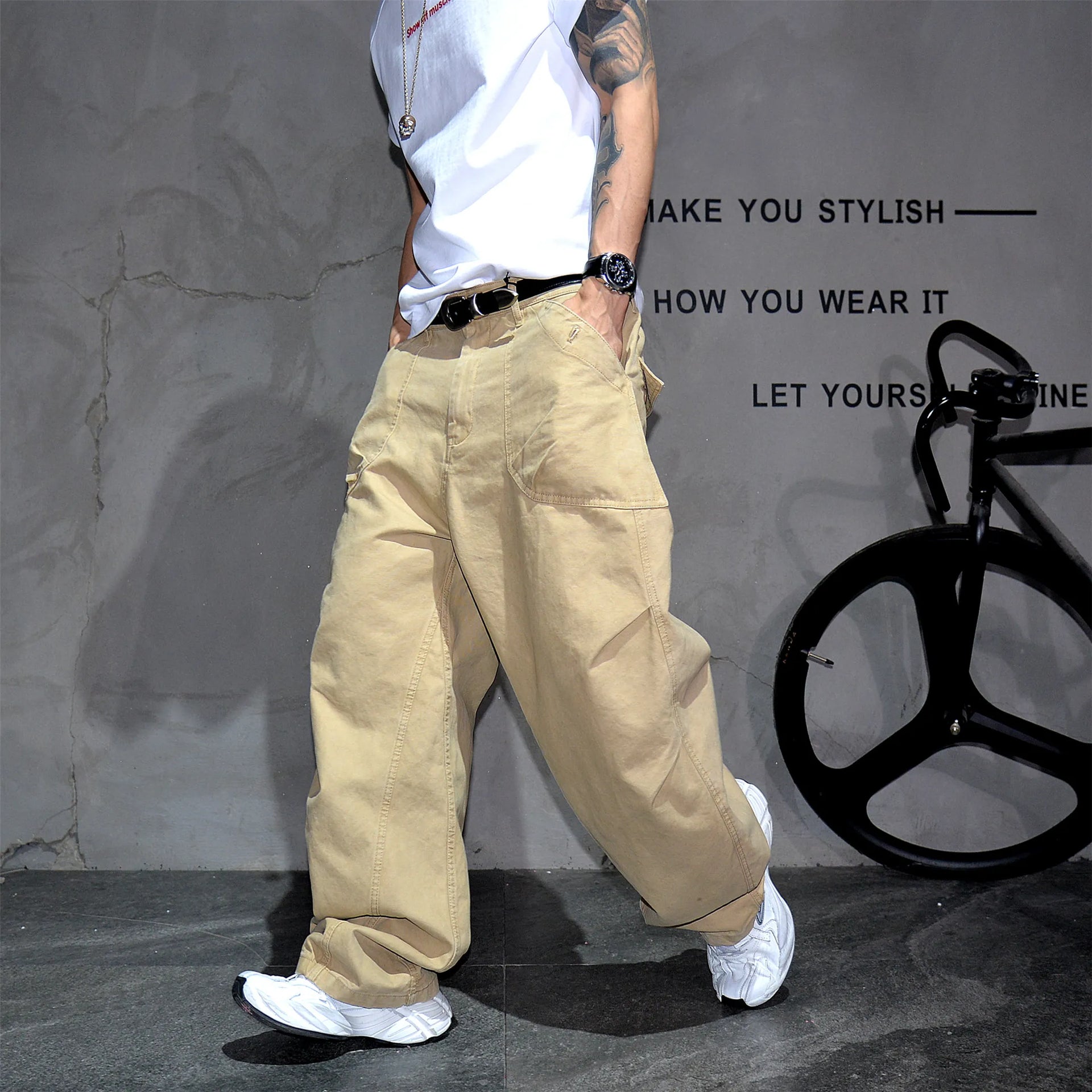 A person wearing loose beige cargo-style pants, a white sleeveless top, and white sneakers stands in a studio setting with a gray textured wall and a black bicycle prop in the background.