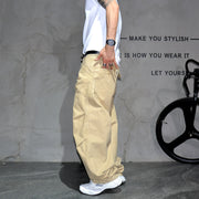 A person wearing loose beige cargo pants, a white t-shirt, and white sneakers stands in front of a textured gray wall with black text and a bicycle wheel visible on the right.