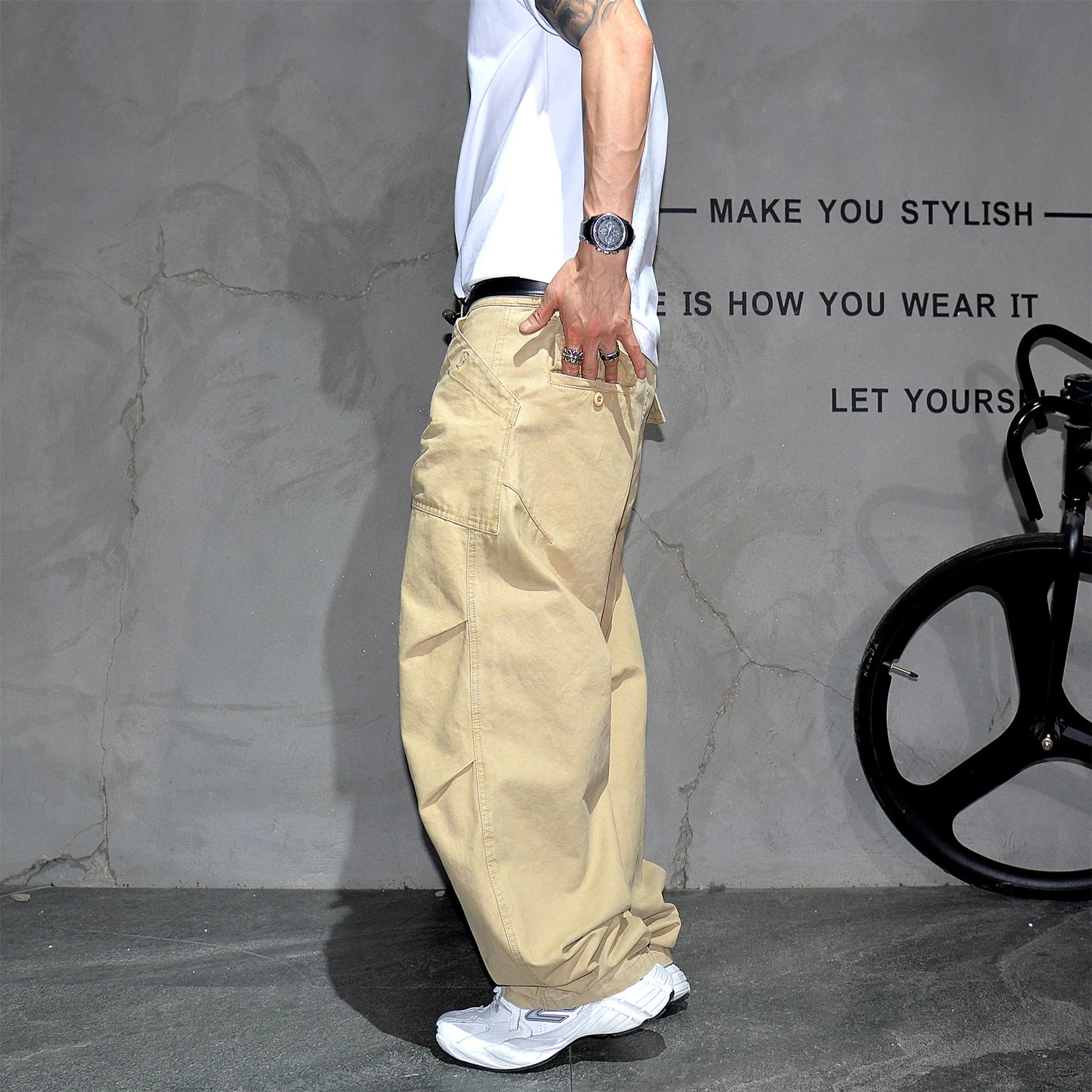 A person wearing loose beige cargo pants, a white t-shirt, and white sneakers stands in front of a textured gray wall with black text and a bicycle wheel visible on the right.