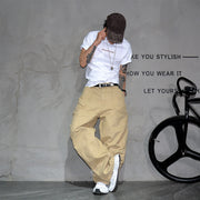 A person wearing a white graphic T-shirt, beige wide-leg cargo pants, a dark cap, and white sneakers poses against a textured gray wall with a black bicycle wheel visible on the right.