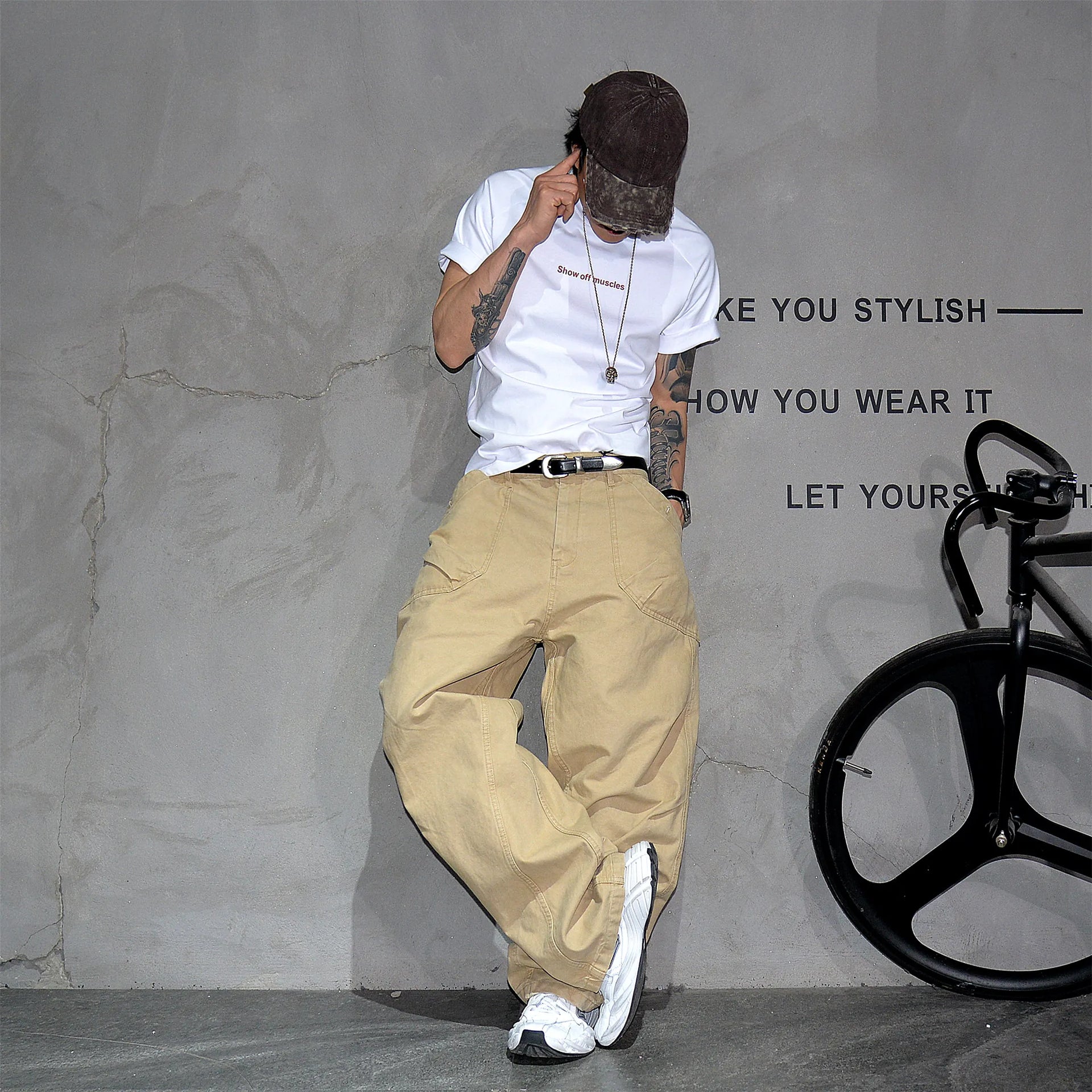 A person wearing a white graphic T-shirt, beige wide-leg cargo pants, a dark cap, and white sneakers poses against a textured gray wall with a black bicycle wheel visible on the right.