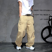 A person wearing baggy beige cargo pants, a white t-shirt, and white sneakers stands against a textured gray wall with black text and a bicycle wheel visible on the right.