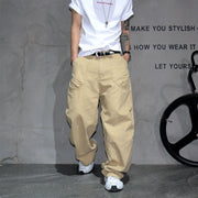A person wearing baggy beige cargo pants, a white t-shirt, and white sneakers, standing against a textured gray wall with black text and a bicycle wheel visible on the right.
