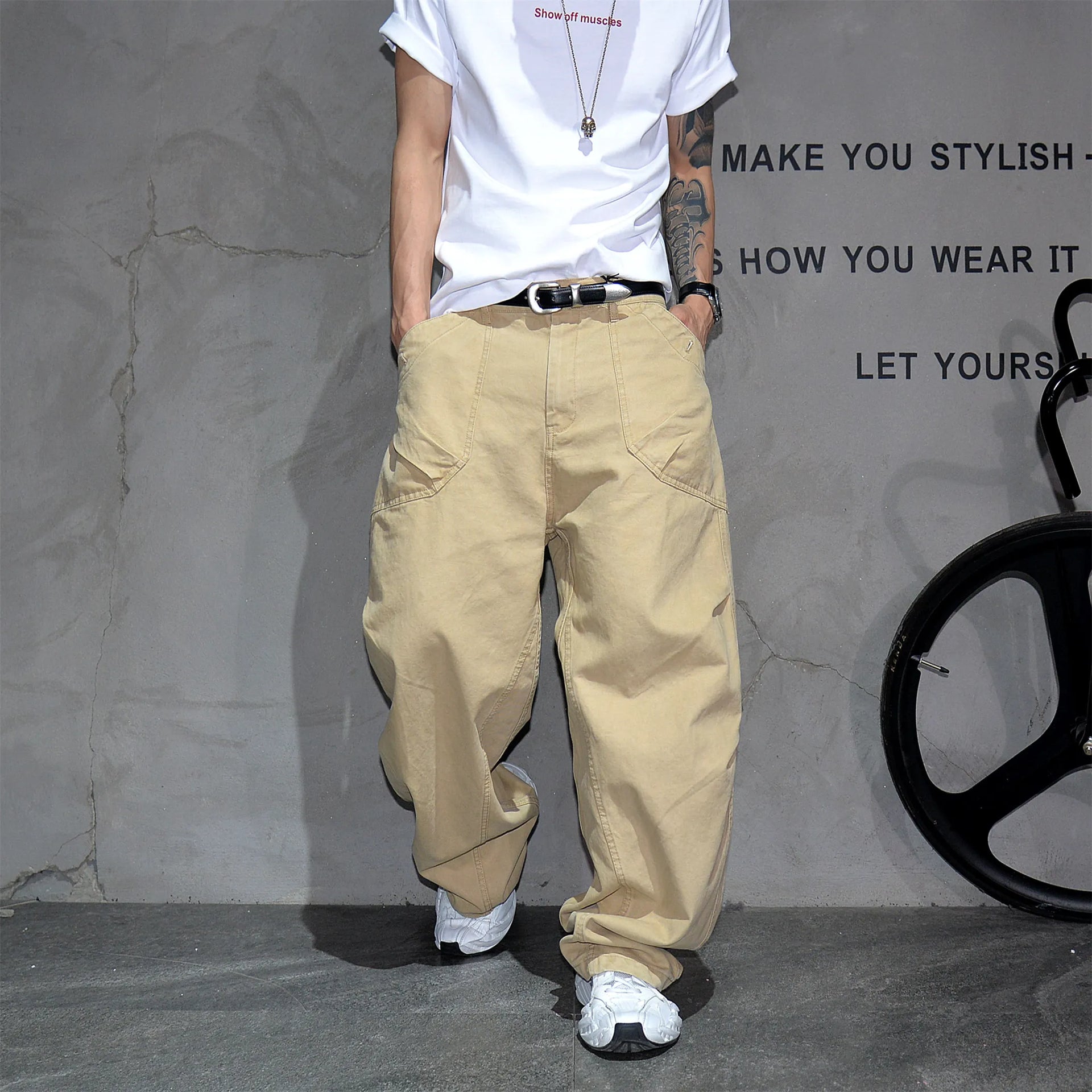 A person wearing baggy beige cargo pants, a white t-shirt, and white sneakers, standing against a textured gray wall with black text and a bicycle wheel visible on the right.