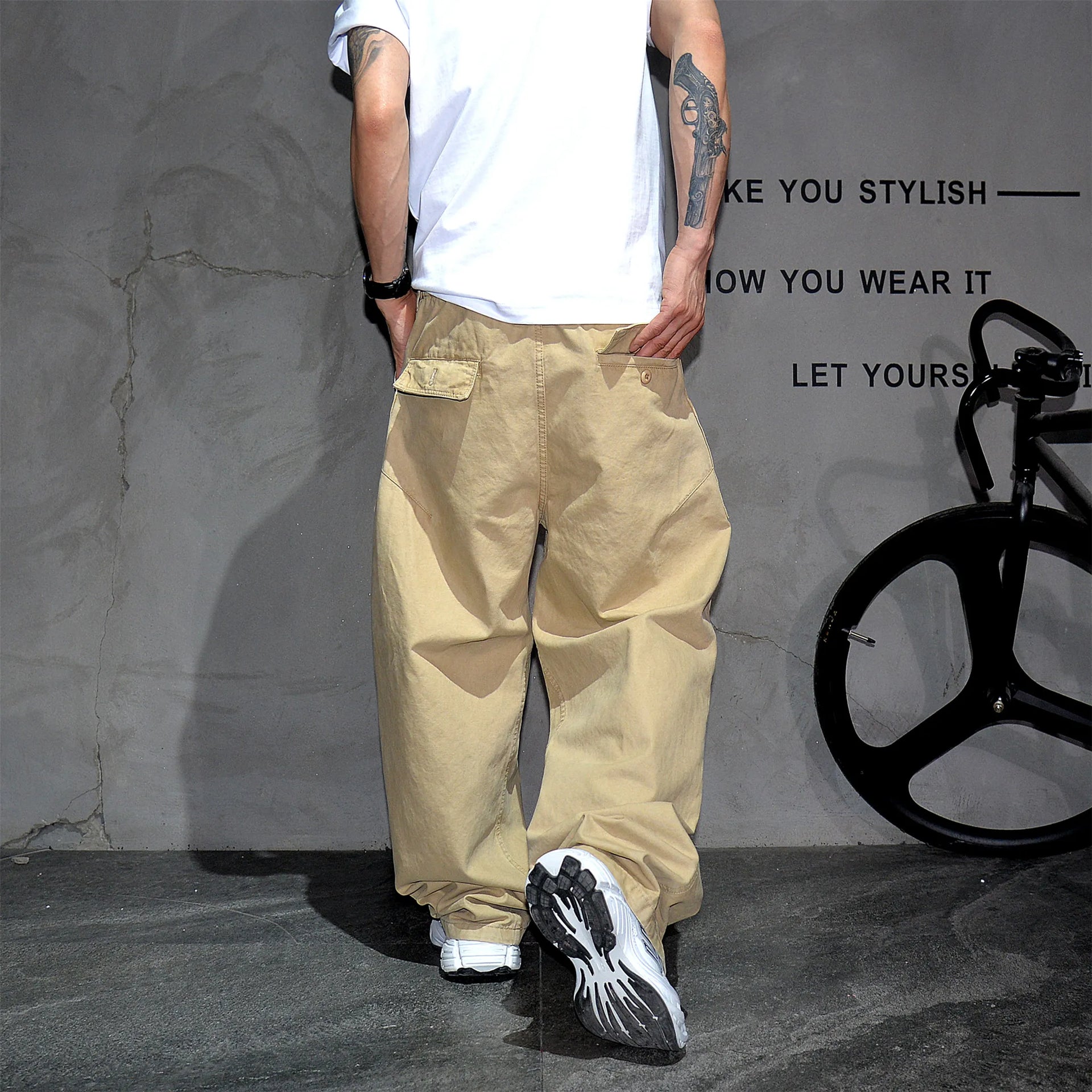 A person from the back wearing baggy beige cargo pants, a white t-shirt, and black-and-white sneakers, standing against a textured gray wall with a black bicycle wheel visible on the right.