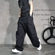 A person wearing loose black wide-leg jeans, a black t-shirt, and white sneakers, standing in front of a textured gray wall with decorative text and a bicycle wheel prop.
