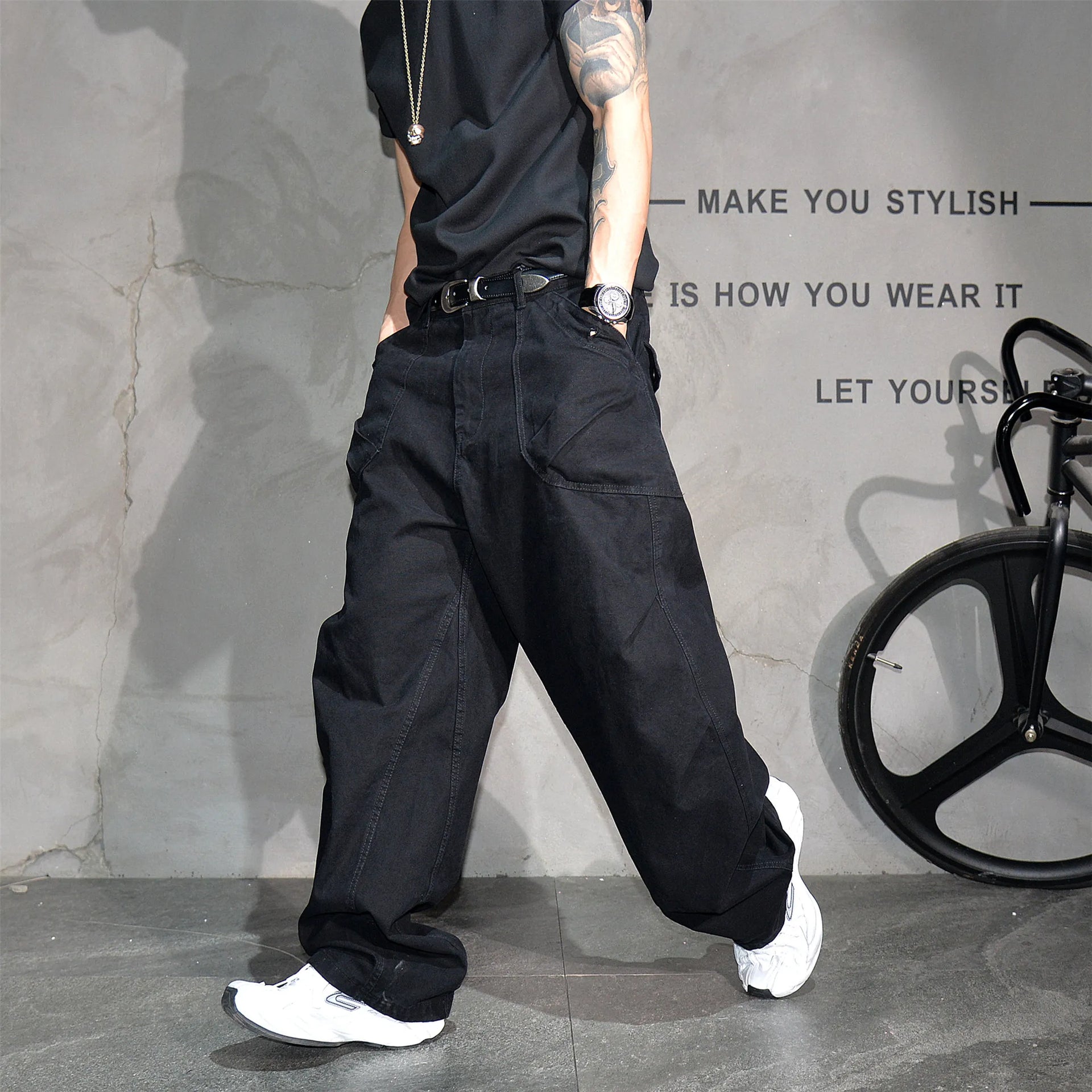 A person wearing loose black wide-leg jeans, a black t-shirt, and white sneakers, standing in front of a textured gray wall with decorative text and a bicycle wheel prop.