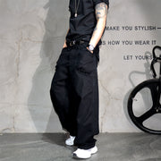 A person wearing loose black wide-leg cargo pants, a black t-shirt, and white sneakers, standing against a textured gray wall with a bicycle wheel visible on the right.