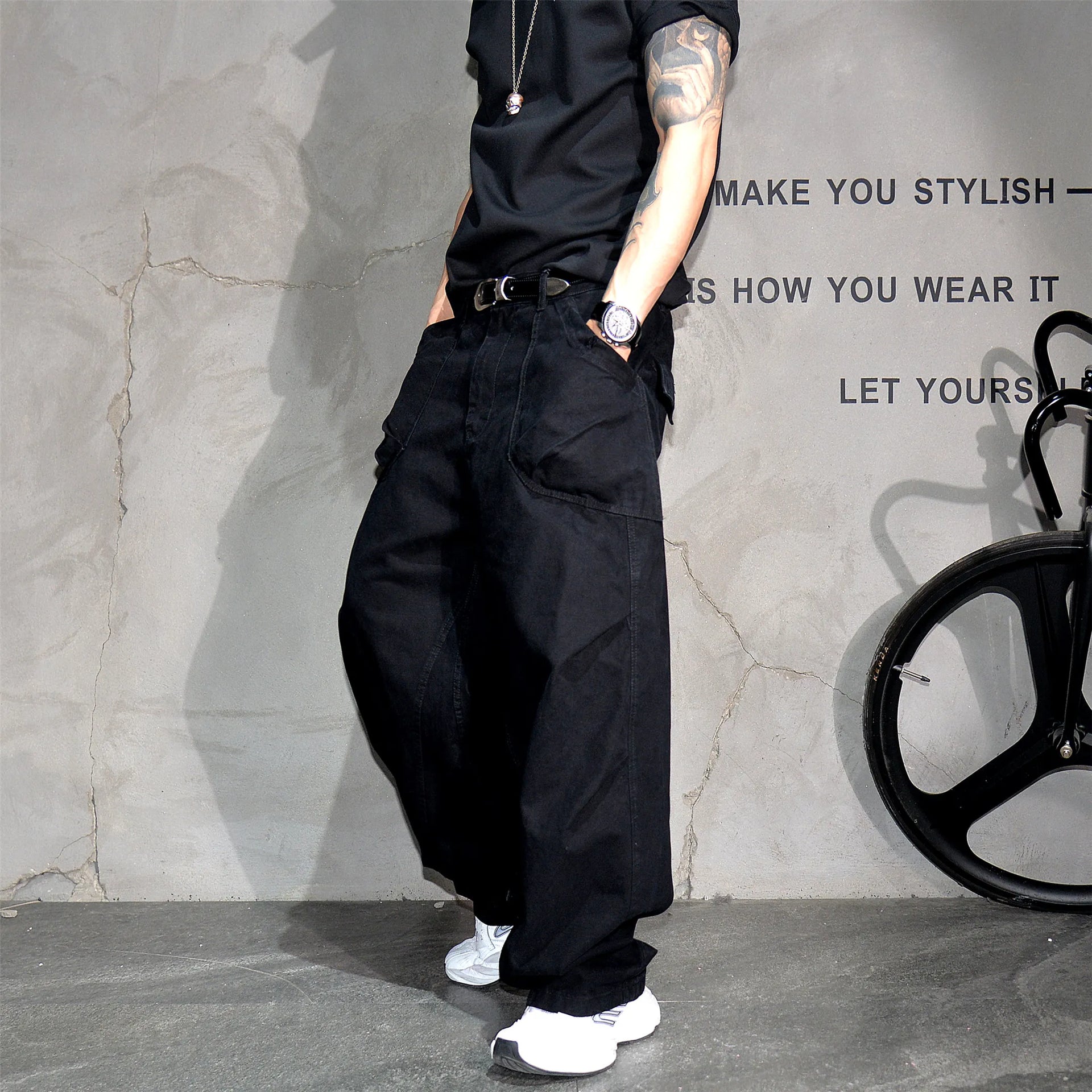 A person wearing loose black wide-leg cargo pants, a black t-shirt, and white sneakers, standing against a textured gray wall with a bicycle wheel visible on the right.