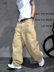 A person wearing loose beige cargo pants, a white t-shirt, a black belt, and white sneakers, standing against a gray wall with black text and a wheel prop.