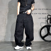 A person wearing loose black wide-leg cargo pants with a black t-shirt, black belt, and white sneakers, standing against a textured gray wall with a bicycle wheel visible on the right.