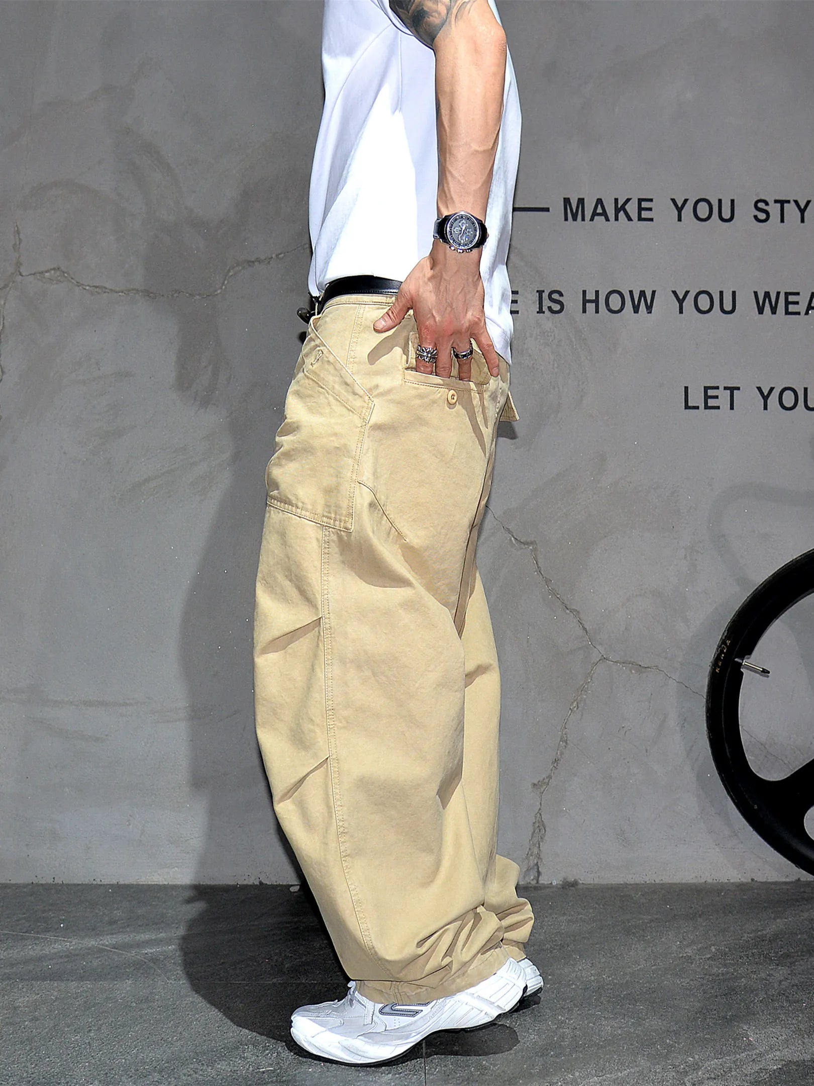 A person wearing loose beige cargo pants, a white t-shirt, and white sneakers, standing against a textured gray wall with black text.