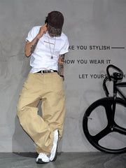 A person wearing a white graphic T-shirt, beige cargo pants, a dark cap, and white sneakers poses against a gray wall with text and a black bicycle wheel visible.