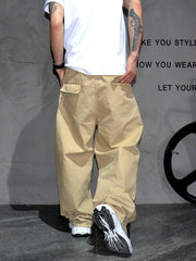 A person shown from the back wearing loose beige cargo-style pants, a white T-shirt, and black-and-white sneakers against a gray wall with partial text and a wheel-like decor element.