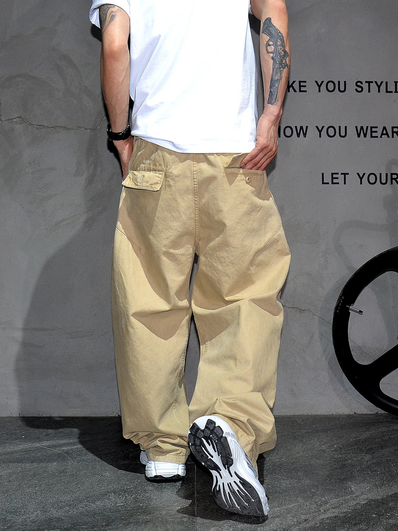 A person shown from the back wearing loose beige cargo-style pants, a white T-shirt, and black-and-white sneakers against a gray wall with partial text and a wheel-like decor element.