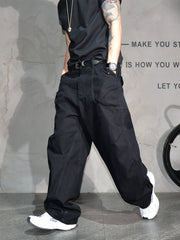 A person wearing loose black wide-leg cargo pants with a black belt, paired with a black t-shirt and white sneakers, standing against a gray textured wall.