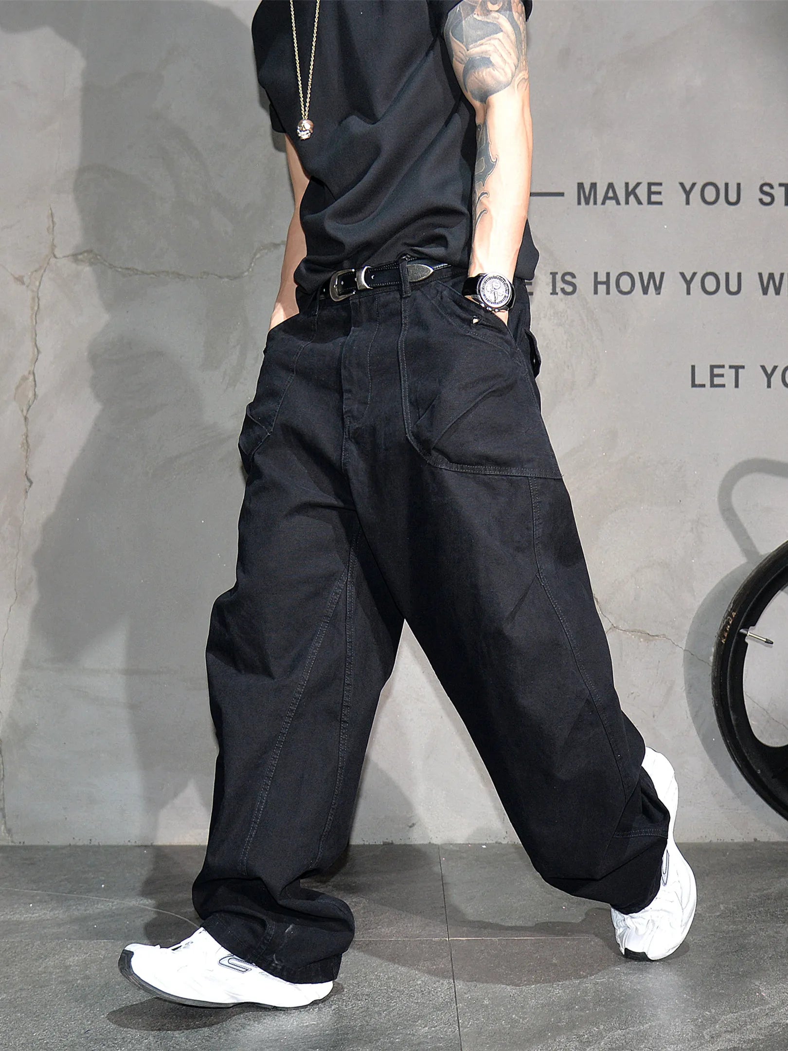 A person wearing loose black wide-leg cargo pants with a black belt, paired with a black t-shirt and white sneakers, standing against a gray textured wall.