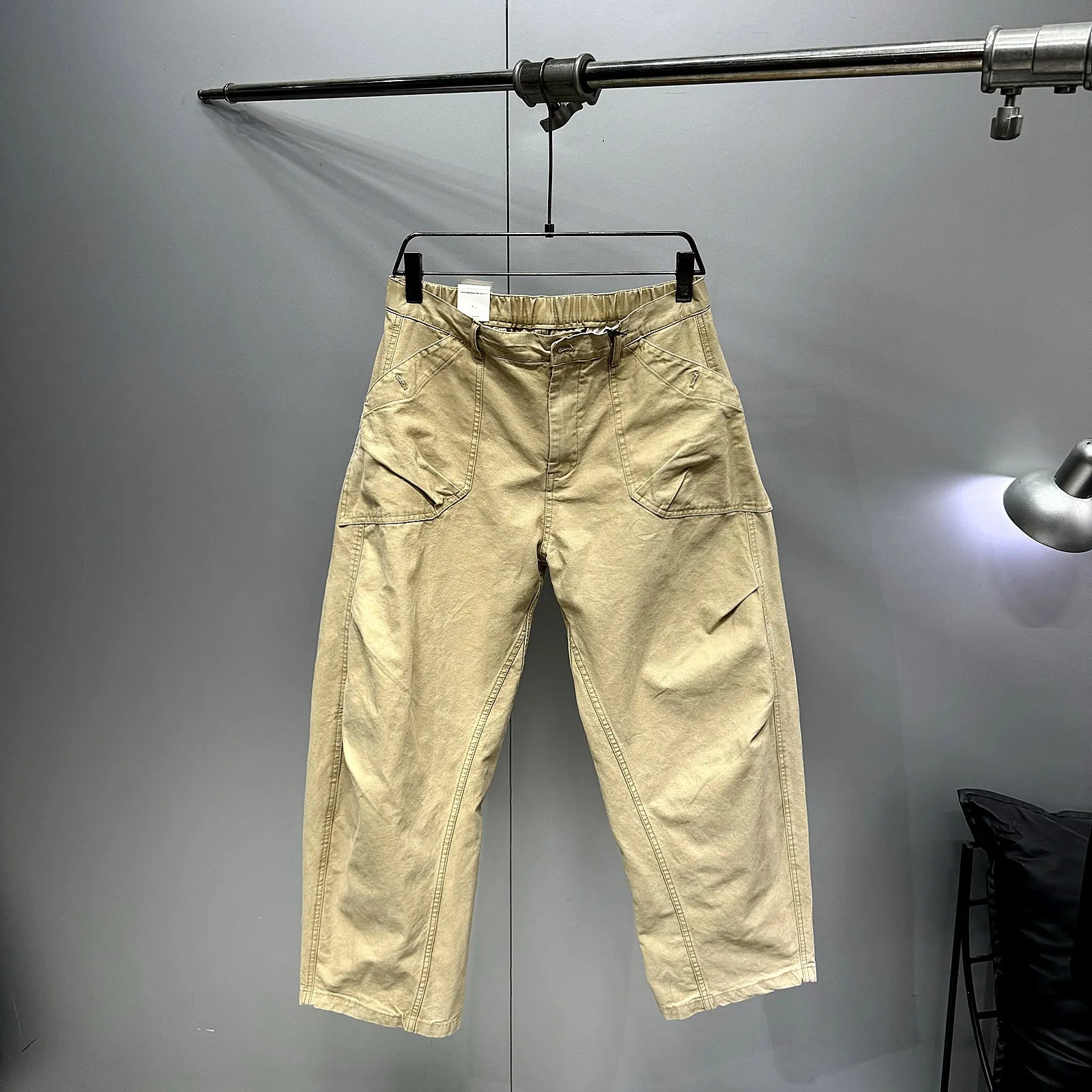 A pair of beige cargo-style pants hanging on a black hanger from a metal clothing rack against a gray wall.