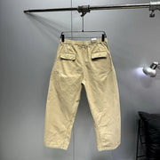 A pair of light beige cropped pants with an elastic waistband and flap back pockets, hanging on a black hanger from a metal clothing rack against a gray wall.
