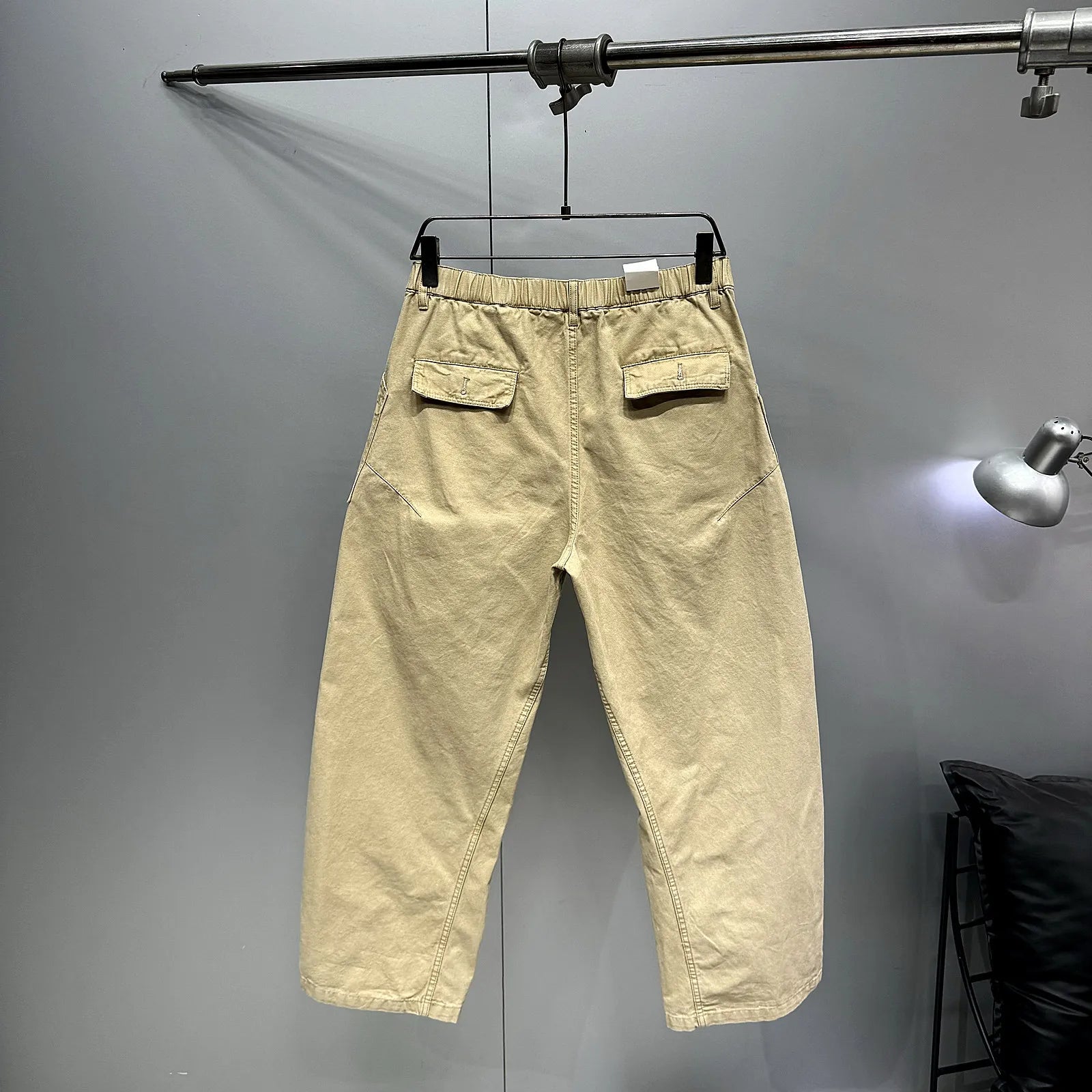 A pair of light beige cropped pants with an elastic waistband and flap back pockets, hanging on a black hanger from a metal clothing rack against a gray wall.