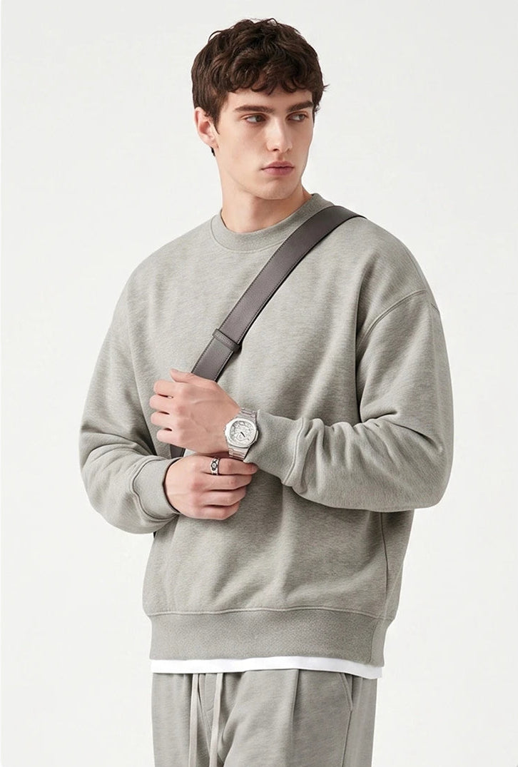 A man wearing a light gray crewneck sweatshirt and matching sweatpants, with a dark gray shoulder strap across his chest, posed against a clean off-white background.