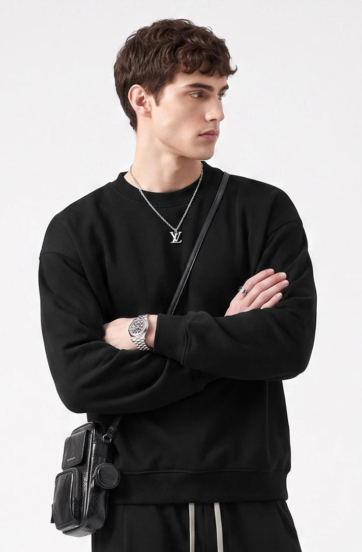 A man wearing a black crewneck sweatshirt, black pants, a silver necklace with a pendant, and a black crossbody bag, posed against a light gray background.