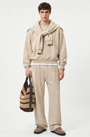 A male model stands against a light gray studio background wearing a beige hooded sweatshirt with a large draped hood, matching beige wide-leg sweatpants, and brown slip-on shoes, while holding a brown and beige striped tote bag.