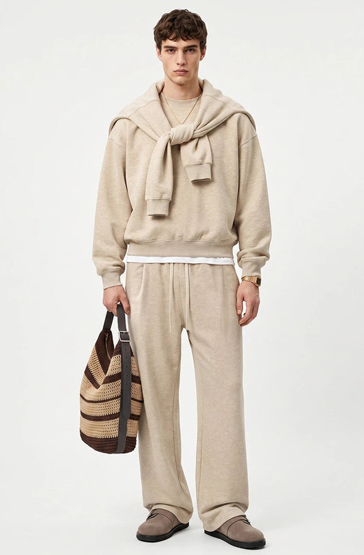 A male model stands against a light gray studio background wearing a beige hooded sweatshirt with a large draped hood, matching beige wide-leg sweatpants, and brown slip-on shoes, while holding a brown and beige striped tote bag.
