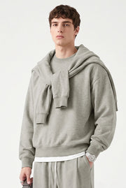 A male model wearing a light gray oversized hoodie with a matching knit scarf draped over the shoulders, paired with matching gray sweatpants, against a clean off-white background.