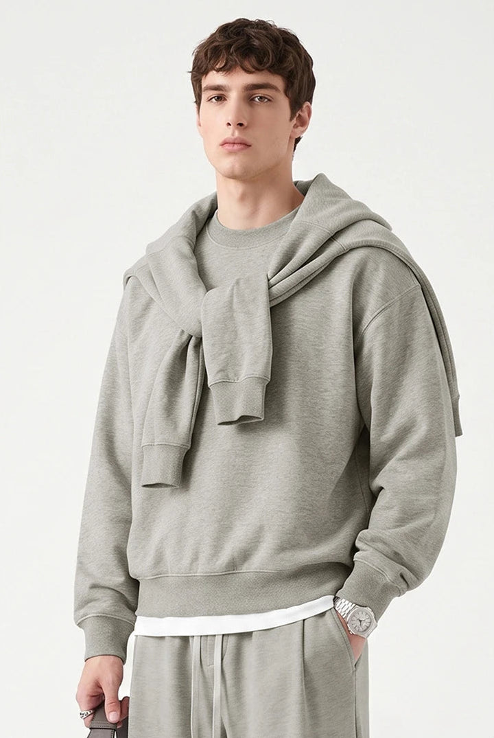A male model wearing a light gray oversized hoodie with a matching knit scarf draped over the shoulders, paired with matching gray sweatpants, against a clean off-white background.