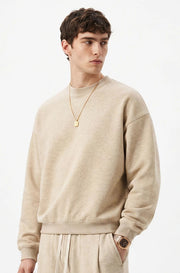 A man wearing a light beige crewneck sweatshirt and matching sweatpants, styled with a gold necklace and watch, set against a plain white background.