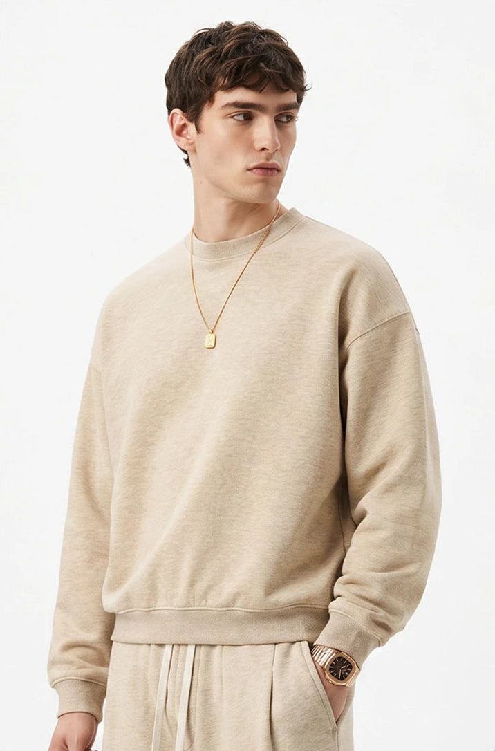 A man wearing a light beige crewneck sweatshirt and matching sweatpants, styled with a gold necklace and watch, set against a plain white background.