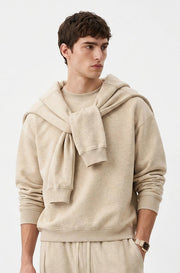 A male model wearing a beige hooded sweatshirt with a large knotted bow detail at the neck, paired with matching beige sweatpants, set against a clean white background.