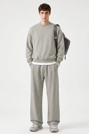 A male model stands against a light gray studio background wearing a light gray crewneck sweatshirt, matching wide-leg sweatpants, and gray sneakers, with a dark gray backpack slung over one shoulder.