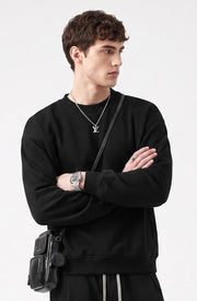 A man wearing a black crewneck sweatshirt, black pants, a silver necklace with a pendant, and a black crossbody bag, posed against a light gray background.