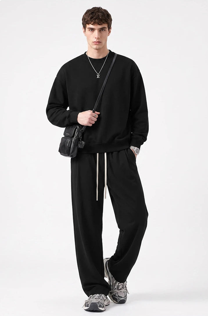 A man modeling a black crewneck sweatshirt and matching black sweatpants with white drawstrings, styled with gray sneakers, a black crossbody bag, and a silver necklace against a light gray background.