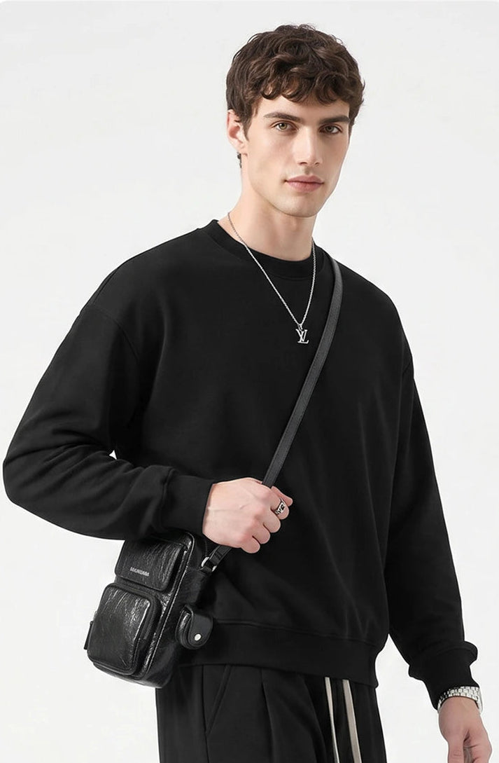 A man wearing a black crewneck sweatshirt and black pants, carrying a black leather crossbody bag with a silver strap. He is wearing a silver necklace with a pendant.