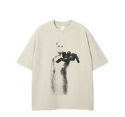 A beige short-sleeve t-shirt featuring a black graphic print of a maskless figure holding a black object, displayed against a solid white background.
